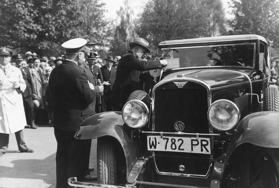 Czarno-białe archiwalne zdjęcie przedstawiające zabytkowy samochód otoczony przez grupę osób podczas wydarzenia motoryzacyjnego. Na pierwszym planie widoczny jest przód klasycznego auta z dużymi reflektorami i charakterystyczną stylistyką z pierwszej połowy XX wieku.